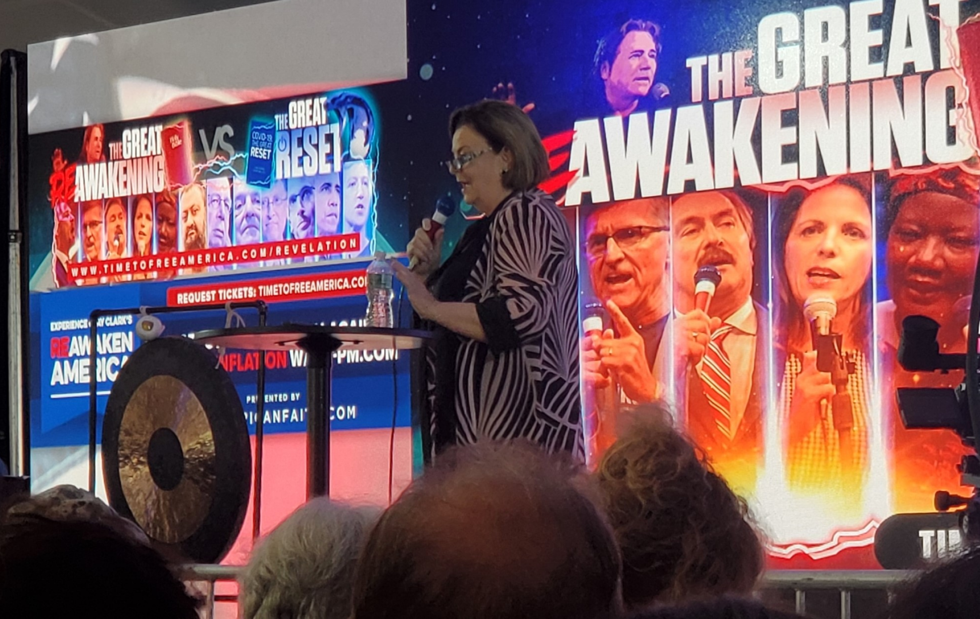 A woman speaks at an event with a large screen behind her displaying political figures.
