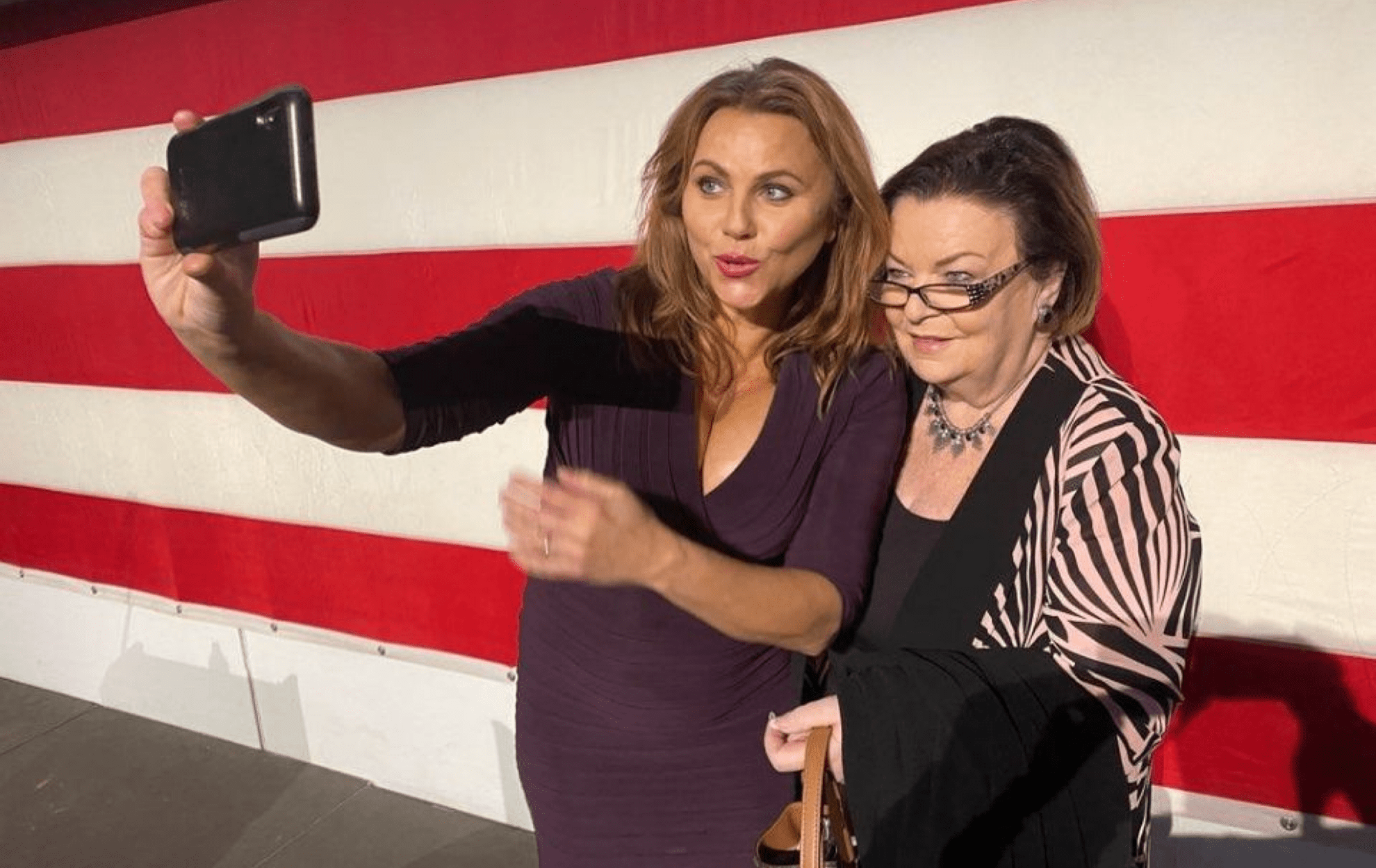 Two women taking a selfie in front of a large American flag.