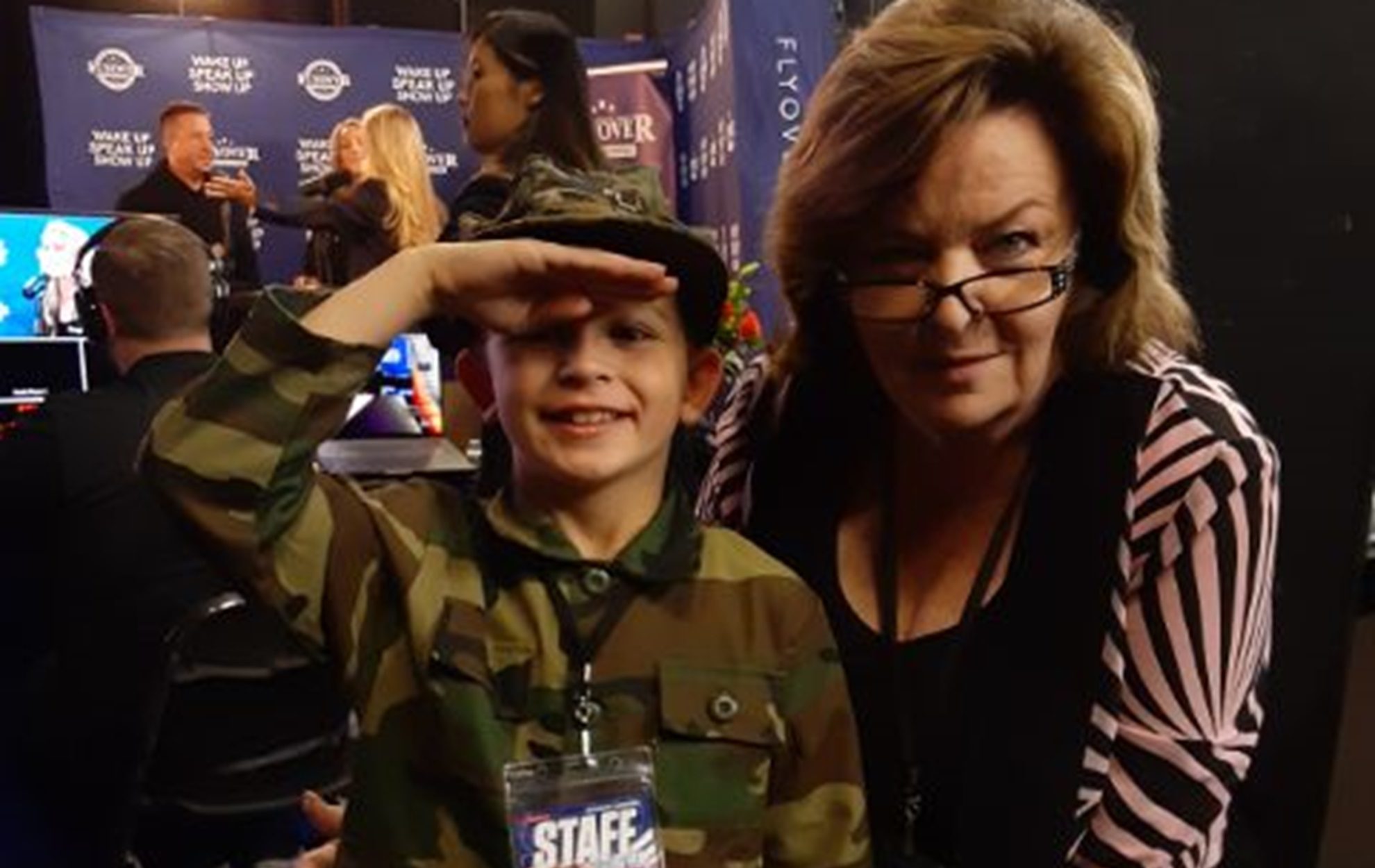 A smiling boy in camouflage salutes next to a woman with glasses indoors.