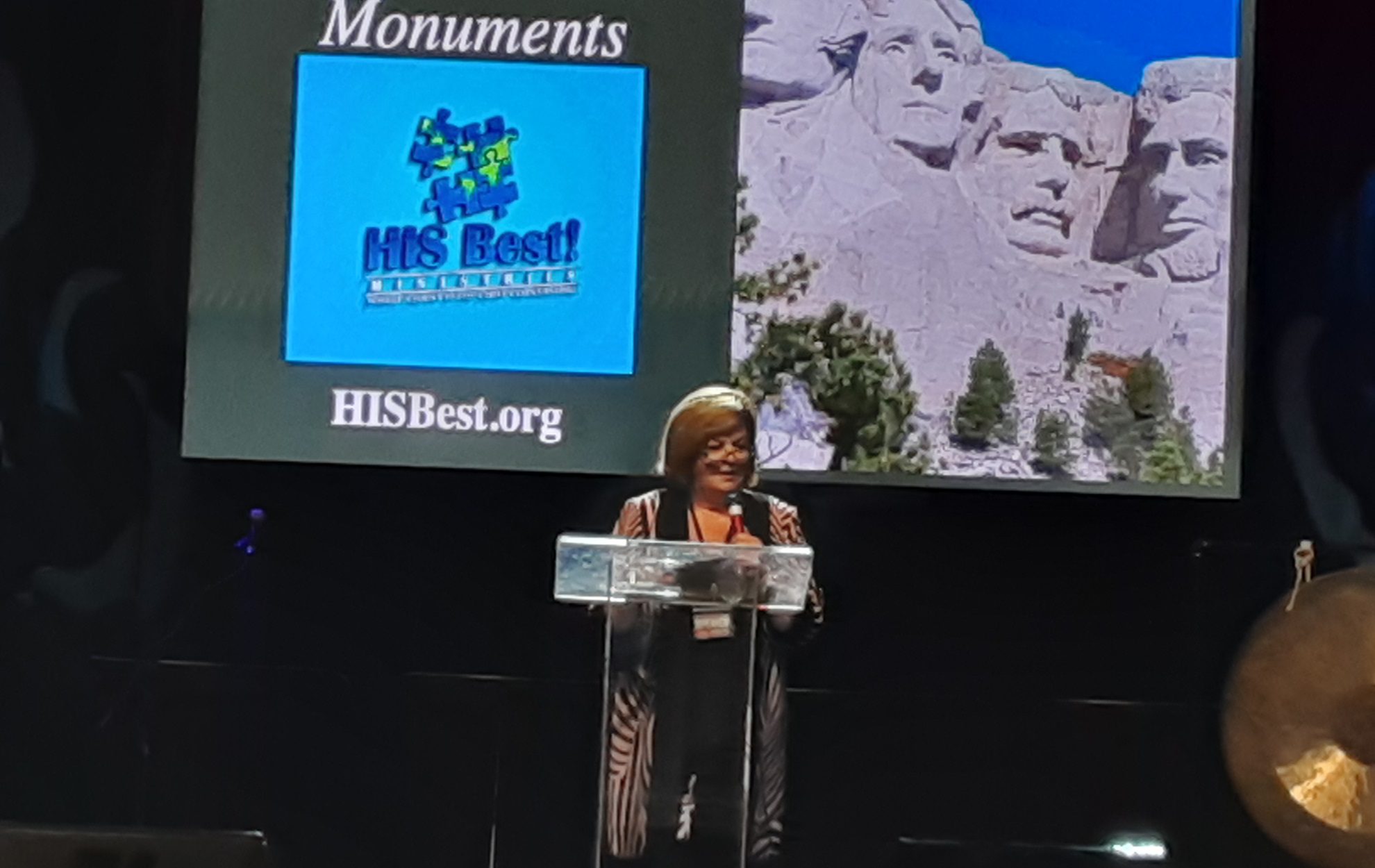 A woman speaks at a podium with Mount Rushmore and a conservation logo projected behind her.