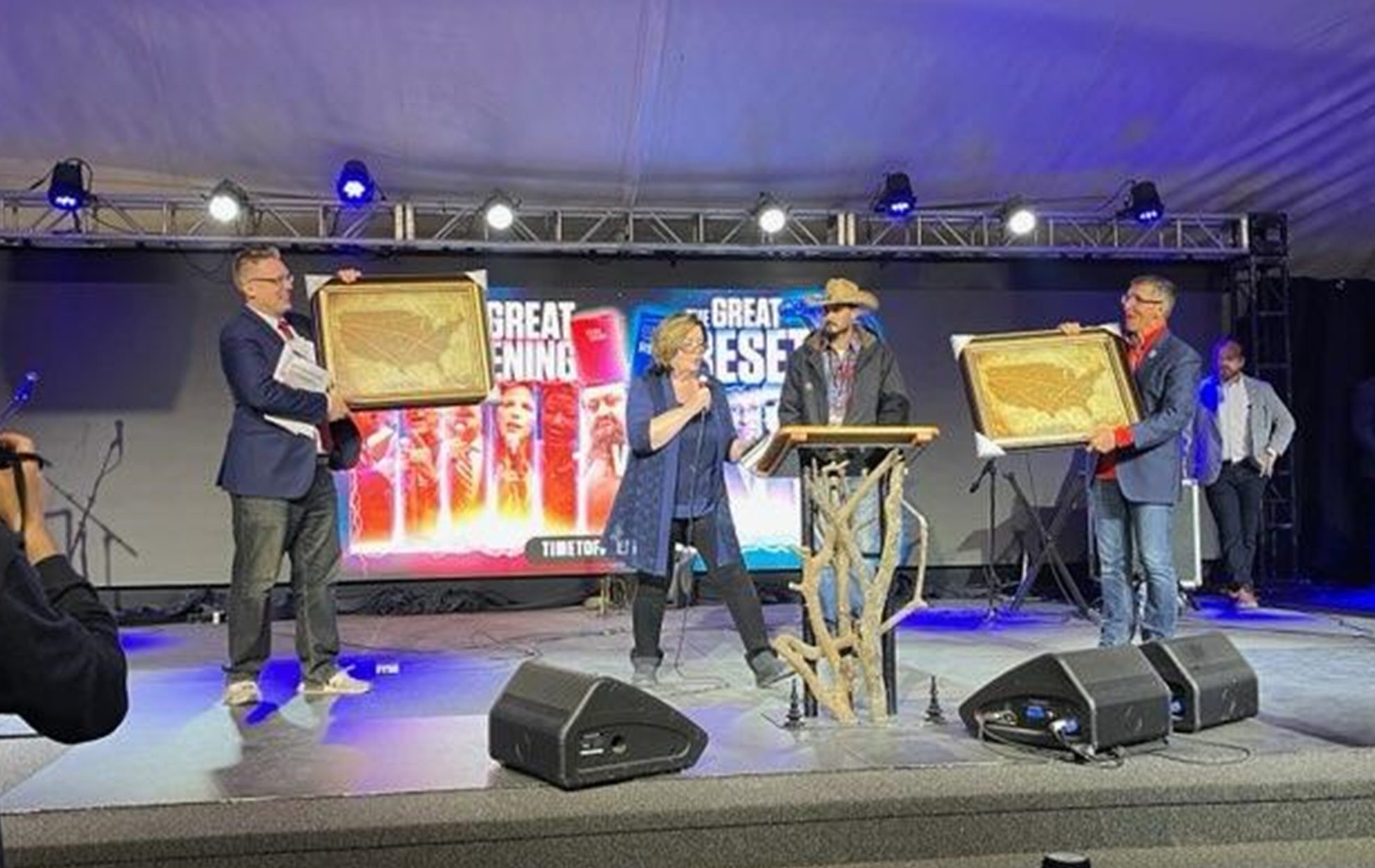 Three people on stage holding awards at an event with a colorful Greatness backdrop.
