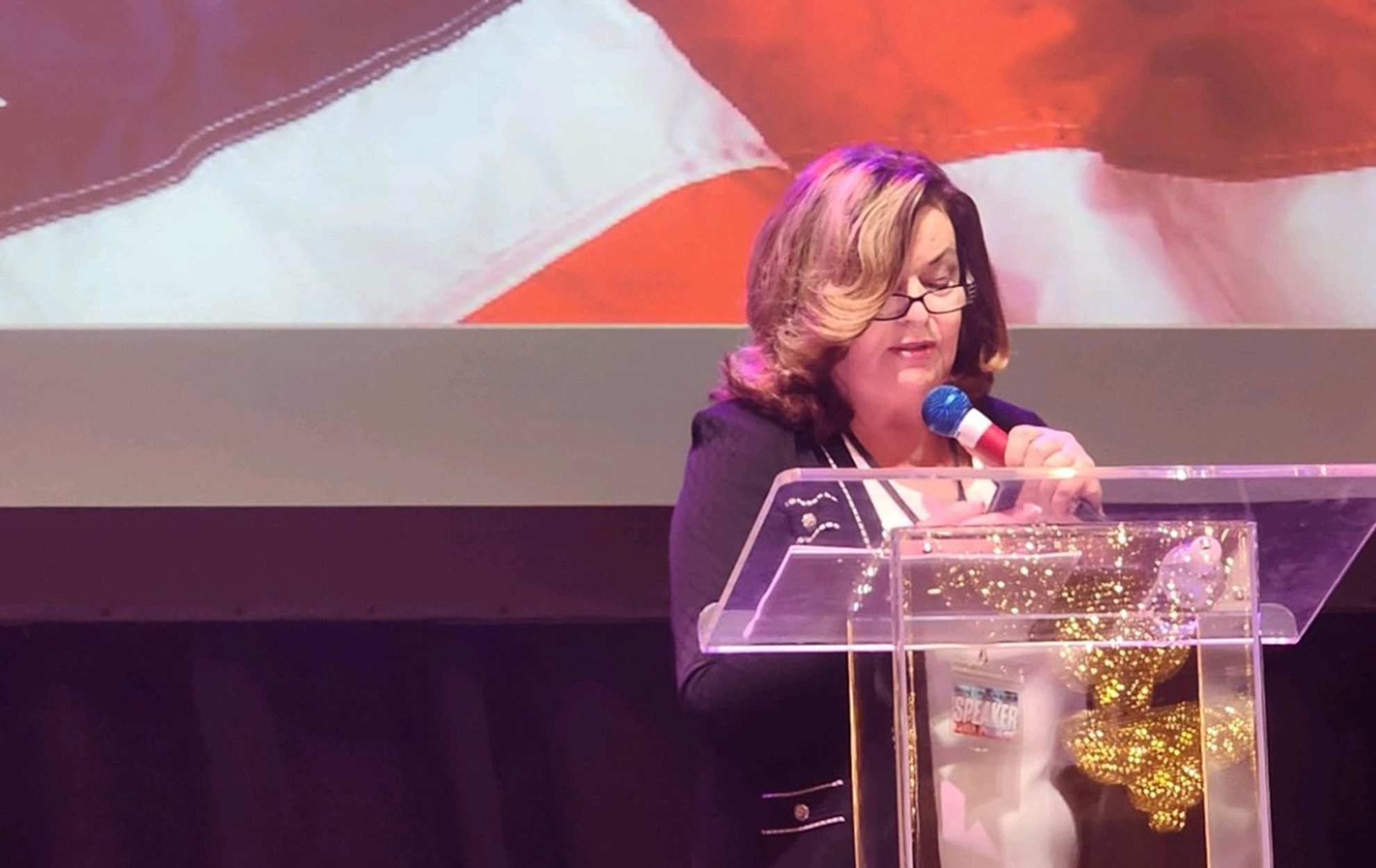 Woman speaking at a podium with a microphone and a large flag in the background.